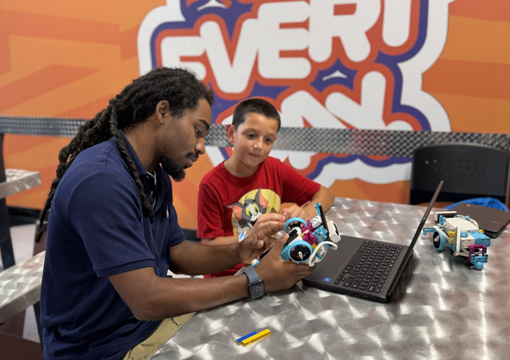 A Code N' Create instructor helping a student build a LEGO SPIKE Prime robot, with an orange "Every Day" graphic behind them